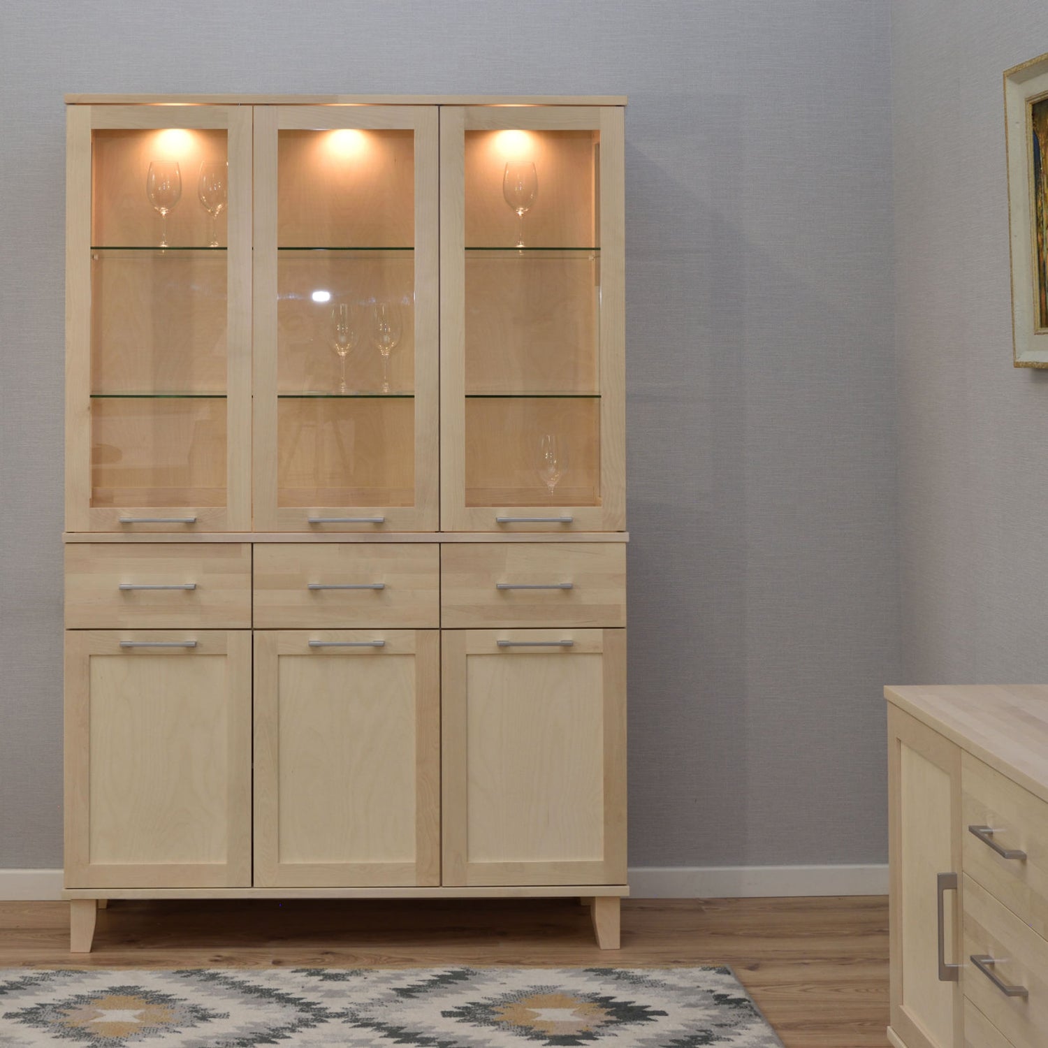 Kuvassa on valkolakattu Vuokatti-vitriini. Vitriinissä on lasikaapisto lasihyllyillä, alaosan tilava senkki kaappien peiliovilla, sekä pitkänmalliset RST-vetimet. Lasikaapin sisällä olevat valot/viinilasit eivät kuulu senkin hintaan.