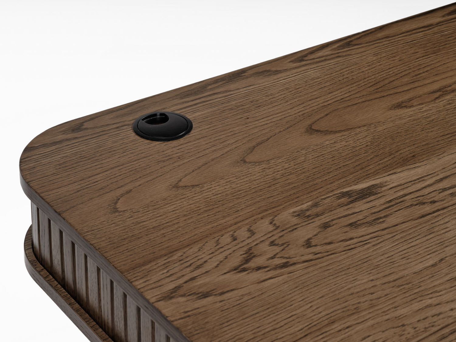 Lähikuvassa Nola-työpöydän kannen pintaa savutammenvärisenä - kannessa on johtoaukko. Koko kalusteen kansilevyn alla olevaa säilytystilaa kiertävät sormipaneloidut liukuovet.