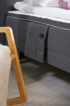 Lähikuvassa antrasiitin värisen Aava-moottorisängyn lisävarusteena saatava tarviketasku. Profiloidussa sijauspatjassa on rungon värinen reunus.