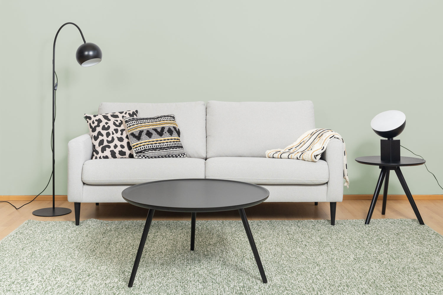 Kuvassa on musta Pod-pöytävalaisin Rowicon Yumi-sarjapöydällä oikealla, sekä vasemmalla musta Round Single-lattiavalaisin. Lisäksi kuvassa on harmaa Mila 3h sohva, vihreä Anna-matto, musta Daisy-sohvapoyta, Hulda ja Rita-koristetyynyt, sekä Sävel Raita-torkkupeitto.