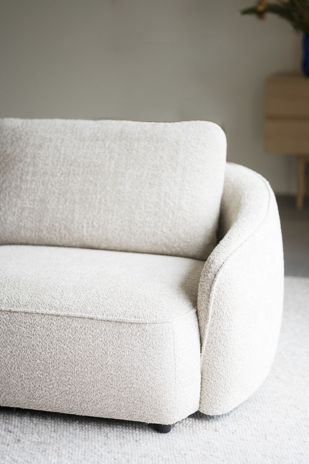 Lähikuvassa Norris-sohvan kaarevaa muotoilua. Verhoiluna vaalea beige Anna 2-kangas ja sohvassa on mustat muovijalat.