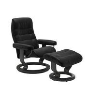 Stressless Opal M -lepotuoli ja rahi mustana: Black 19 Batick-nahkaverhoilulla ja harmaa petsatulla -Classic-puujalustalla.