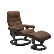 Stressless Opal M -lepotuoli ja rahi ruskeana: Warm Brown 87 Batick-nahkaverhoilulla ja harmaa petsatulla -Classic-puujalustalla.