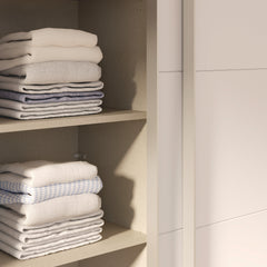 Kuvassa on mattavalkoinen Kira-liukuovikomero - toinen liukuovi avoinna ja kaappia on täydennetty näkyvissä olevilla erikseen myytävillä lisähyllyillä. Kaapin sisus on väriltään harmaata kangasjäljitelmää.