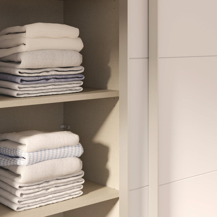 Kuvassa on mattavalkoinen Kira-liukuovikomero - toinen liukuovi avoinna ja kaappia on täydennetty näkyvissä olevilla erikseen myytävillä lisähyllyillä. Kaapin sisus on väriltään harmaata kangasjäljitelmää.