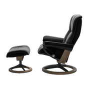 Stressless Mayfair M -lepotuoli ja rahi mustana: Black 19 Paloma-nahkaverhoilulla ja savutammi 13 petsattu pyökki/mattamusta 40 metalli -Signature-yhdistelmäjalustalla.