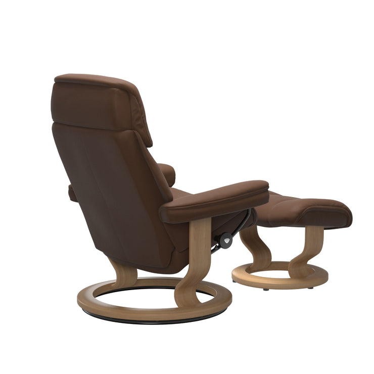Stressless Ruby M -lepotuoli ja rahi ruskeana: Warm Brown 87 Batick-nahkaverhoilulla ja tammi petsatulla -Classic-puujalustalla.