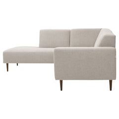 Vasemmanpuoleinen divaallinen Tessa-avokulmasohva vaalealla hiekanvärisellä Taste 13 -kankaalla, 20 cm leveällä käsinojalla ja pähkinänvärisillä 18 cm pyöreäkartiopuujaloilla.