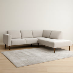 Oikeanpuoleinen divaallinen Tessa-avokulmasohva vaalealla hiekanvärisellä Taste 13 -kankaalla, 20 cm leveällä käsinojalla ja pähkinänvärisillä 18 cm pyöreäkartiopuujaloilla. Stailattu visualisointi.