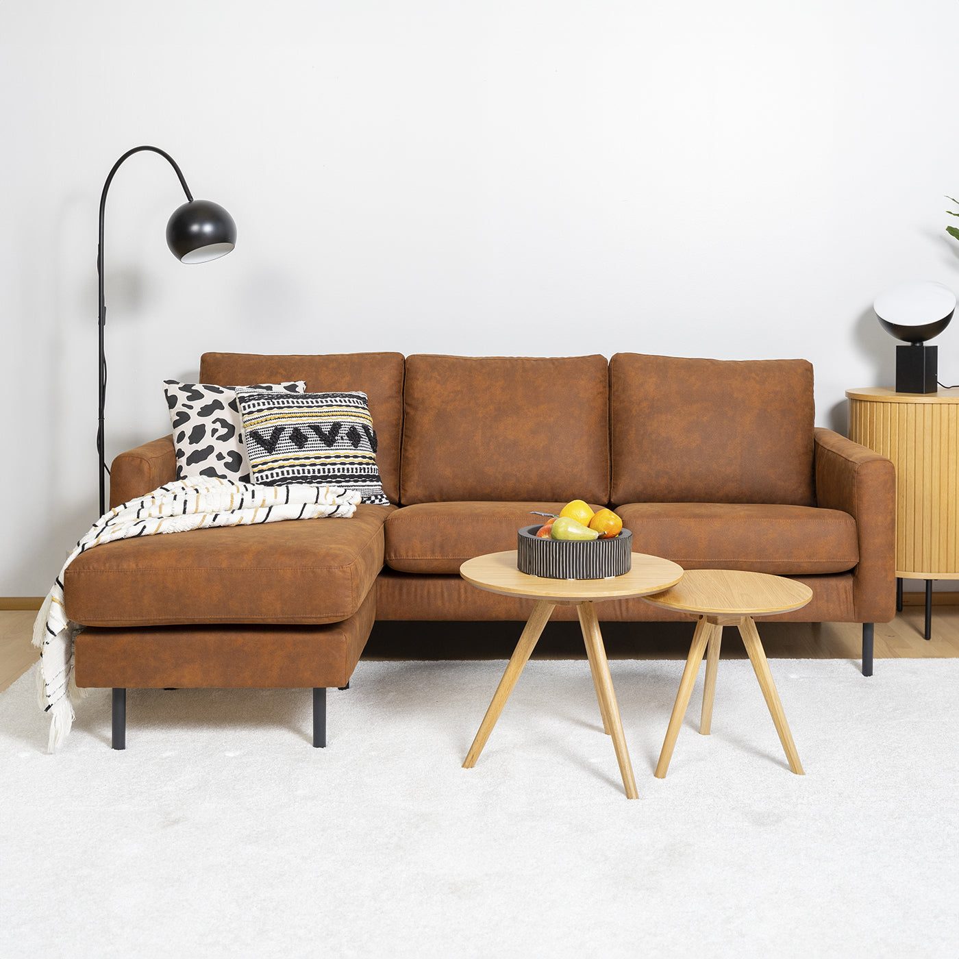 Kuvassa on kolmen istuttava Mila-divaanisohva (divaanin kätisyys vaihdettavissa) joka on verhoiltu ruskealla Texas-kankaalla ja siinä on metalliset ohuet mustat pyöreät tolppajalat. Kuvassa on myös Rowicon Yumi-sarjapöytä ja valaisimet Pod-pöytä- ja Round Single-lattiavalaisin. Oikeassa reunassa on näkyvissä Nola-senkkiä. Mattokympin sisustustuotteet: Louna-matto, Rita & Hulda-koristetyynyt ja Sävel Raita -torkkupeitto.